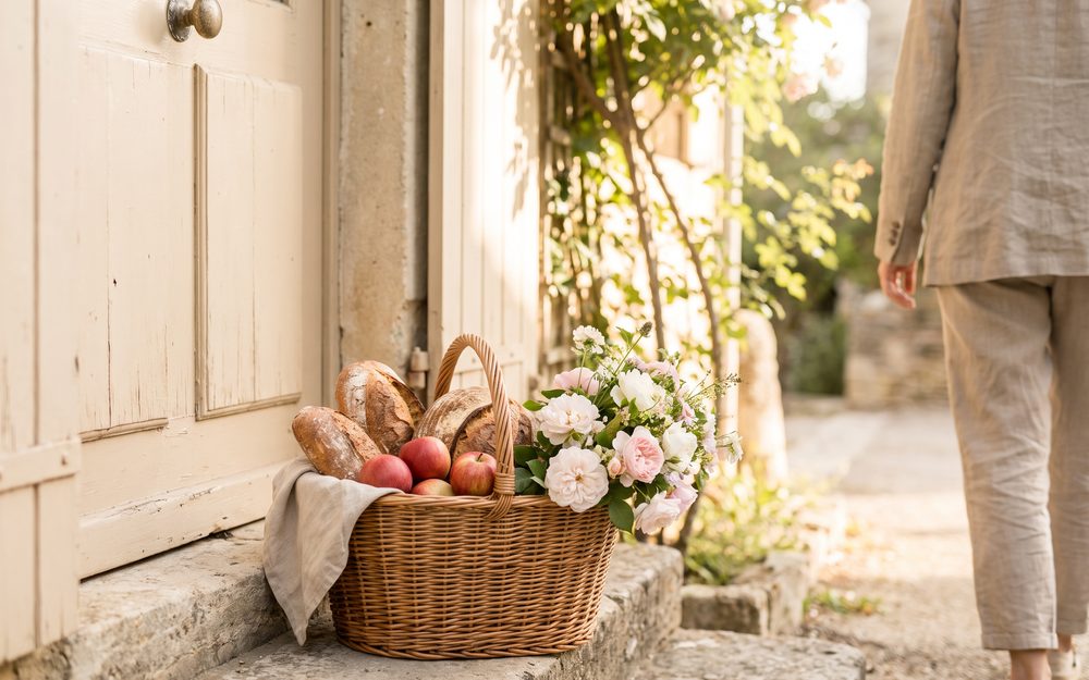 Einkaufskorb mit Brot, Äpfeln und Blumen vor einer hellen Haustür